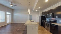 Located at 4906 Wurzbach Pkwy in San Antonio, Texas, this single-floor residence offers a contemporary design with a cohesive, open-concept layout. The living area is generously proportioned, featuring durable wood-style flooring, neutral-toned walls, and ceiling fans that contribute to a clean, modern aesthetic. Natural light filters in through multiple windows fitted with blinds, enhancing the bright and airy atmosphere.

The kitchen serves as a central focal point, anchored by a large island with granite countertop and an integrated sink, illuminated by stylish pendant lighting overhead. Dark espresso cabinetry extends to the ceiling, providing ample storage, while a full suite of stainless steel appliances—including a refrigerator, electric range, and microwave—adds a sleek, functional finish. A tiled backsplash completes the polished look.

The bedroom is carpeted for comfort and receives natural light through a pair of well-positioned windows. The bathroom features wood-style flooring, a granite-topped vanity with dark cabinetry, a large mirror, and a private water closet. Recessed lighting throughout the residence ensures consistent, well-distributed illumination across all areas.