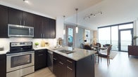 This contemporary single-floor residence showcases a thoughtfully designed open-concept layout that seamlessly connects the kitchen, dining, and living areas. The chef's kitchen is a standout feature, boasting rich dark cabinetry, light granite countertops, a large center island with a double undermount sink, stainless steel appliances including a side-by-side refrigerator, built-in microwave, and range, all complemented by a clean tile backsplash and light hardwood-style flooring. Elegant pendant lighting and a track lighting system add warmth and functionality to the space. The living area is bathed in natural light through expansive floor-to-ceiling windows and a glass door that opens to a private balcony, offering sweeping panoramic views of the surrounding cityscape and greenery. The bedroom continues the refined aesthetic with large windows that frame picturesque outdoor views, light hardwood flooring, and a calm, neutral color palette. The en-suite bathroom features granite countertops, dark cabinetry, a glass-enclosed shower, and a large soaking tub, with direct access to a spacious walk-in closet. Throughout, the residence maintains a cohesive modern style with soft gray tones, quality finishes, and an abundance of natural light.