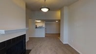 This single-floor residence offers an open and functional layout that connects the living and kitchen areas seamlessly. The living room serves as the heart of the space, anchored by a classic fireplace with black tile surround and a white mantel, adding a traditional focal point to the room. Neutral-toned carpeting extends throughout the living and adjoining areas, contributing to a warm and cohesive atmosphere.

A pendant light fixture hangs from the ceiling, providing ambient illumination over the main living area. The kitchen is positioned adjacent to the living room and is accessible via a pass-through counter, which enhances the open feel of the layout while maintaining a defined cooking zone. White cabinetry lines the kitchen walls, complemented by overhead lighting that brightens the workspace. A range and refrigerator are visible within the kitchen.

Beyond the kitchen, an additional open area provides flexible space that could serve multiple purposes. The overall color palette throughout the residence is neutral, with beige and white tones dominating the walls and flooring, creating a clean and understated aesthetic well-suited for personalization.