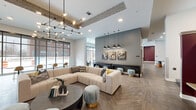 Located at 419 Donald Driver Way in Green Bay, Wisconsin, this thoughtfully designed single-floor community space offers a generous and versatile layout ideal for gathering and socializing. The open-plan living area is anchored by a large, plush sectional sofa set atop striking herringbone-patterned hardwood flooring, complemented by an eye-catching geometric pendant chandelier and exposed concrete ceiling beams that add an industrial-modern character. A bold accent wall in deep burgundy provides a dramatic focal point, while soft gray tones throughout the space create a cohesive, contemporary palette. The dining area features a round table with white Windsor chairs beneath elegant globe pendant lights, framed by a richly patterned feature wall with built-in shelving and a wall-mounted display screen. Floor-to-ceiling windows fitted with roller shades flood the interior with natural light and offer views of the surrounding streetscape. A wide corridor connects various seating zones, each thoughtfully appointed with lounge chairs, bistro tables, and casual bar-height seating along the window line. Textured accent columns and layered artwork add depth and personality throughout. The overall ambiance is polished yet welcoming, blending upscale residential comfort with functional communal design.