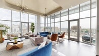 Located at 517 N Shipley Street in the heart of downtown Wilmington, Delaware, this elevated lounge space delivers a striking blend of modern architecture and refined interior design. The defining feature of the space is its dramatic floor-to-ceiling curtain wall glazing, which wraps the perimeter and floods the interior with abundant natural light while offering expansive panoramic views of the Wilmington skyline.

The open-plan layout is divided into distinct seating zones, each thoughtfully curated with contemporary mid-century modern furnishings. A bold, curved blue velvet sofa serves as a focal point in one area, complemented by warm cognac leather armchairs and neutral upholstered seating arrangements. A sleek linear fireplace is set into a textured accent wall, adding warmth and architectural interest alongside a large flat-screen display and a built-in shelving unit styled with decorative objects and lush greenery.

Large-format tile flooring and layered area rugs define each zone, while statement pendant chandeliers and floor lamps provide layered ambient lighting. Glass-partitioned entry vestibules and interior glass doors maintain visual openness throughout. Vibrant fiddle-leaf fig trees and potted palms introduce organic texture, enhancing the space's inviting and sophisticated atmosphere.