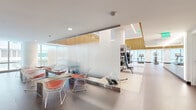 Located at 33 8th Street in San Francisco's SoMa neighborhood, this contemporary fitness center offers a thoughtfully designed, single-floor layout that accommodates a full range of workout needs. The space is defined by its striking architectural elements, including warm vertical wood slat panels lining the upper walls and ceiling perimeter, crisp white surfaces, and floor-to-ceiling windows that flood the interior with natural light and frame views of the surrounding cityscape.

The cardio zone features a generous row of treadmills, elliptical trainers, stationary bikes, and rowing machines, all positioned to take advantage of the expansive window views. The strength training area is well-equipped with a multi-functional cable and weight rack system, free weights, medicine balls, and a dedicated stretching and functional training zone with padded flooring. Full-length mirrors along one wall visually expand the space and support form-focused training.

A frosted glass partition elegantly divides a flexible lounge or stretch area furnished with modern wire-frame chairs, adding a stylish, multifunctional element to the layout. A sleek kitchenette with bold red cabinetry and a built-in sink provides added convenience, complemented by nearby vending machines. The overall ambiance is clean, energizing, and modern, reflecting a high-quality fitness environment.