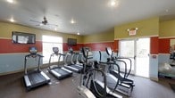 This single-floor fitness center is designed to provide a practical and motivating environment for cardio-focused exercise. The space is outfitted with a row of treadmills and elliptical machines from True Fitness, offering users a solid range of cardio equipment to choose from. The walls feature a bold tri-color scheme — olive green on the upper portion, deep red as an accent band, and light grey along the lower section — creating a dynamic and energizing atmosphere. A ceiling fan centered overhead aids in air circulation and comfort during workouts, while recessed lighting and a wall-mounted flat-screen television add to the functional, gym-like environment. Natural light fills the room through multiple windows, including a large window on the far wall and windows along the side, contributing to an open and airy feel. Durable, textured flooring is well-suited for heavy equipment and high-traffic use. An illuminated exit sign and exterior door provide convenient access and safety compliance. The overall layout is clean and organized, making efficient use of the available space to accommodate multiple users simultaneously.