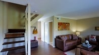 This two-story residence offers a well-organized and comfortable layout spread across two thoughtfully designed floors. The main level showcases rich hardwood flooring that flows seamlessly throughout the space, lending warmth and a sense of continuity. The living area is welcoming and functional, anchored by a plush leather sofa accented with decorative pillows and complemented by warm wall art in earthy tones, adding a vibrant focal point to the room. Crown molding lines the walls, contributing a refined architectural detail to the interior. A staircase with classic white balusters provides an elegant transition to the upper level, while also serving as a natural divider between the living and entryway areas. Near the base of the stairs, a convenient entryway nook offers practical storage space. Closet storage is readily accessible on the main floor, adding to the home's functionality. Ceiling vents throughout suggest a central HVAC system, ensuring year-round comfort. The textured ceiling adds subtle character overhead. Overall, the space strikes a balance between casual comfort and understated style, making it a practical and pleasant environment suitable for everyday living.