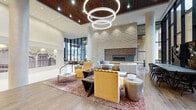 This thoughtfully designed lobby makes an immediate impression with its soaring double-height ceilings and expansive floor-to-ceiling windows that flood the space with natural light. Suspended overhead, a cascading trio of circular LED chandeliers serves as a bold focal point, complemented by a slatted wood ceiling detail that adds warmth and texture.

The lounge area is anchored by a full-height stacked stone accent wall housing a sleek linear fireplace, framed by plush white sofas, velvet accent chairs in warm gold tones, and a richly patterned area rug. Metallic decorative accessories and a geometric brass coffee table add layers of visual interest.

A sculptural elephant artwork stands prominently near the entry, adding a distinctive artistic character to the space. The entry itself features a set of black-framed glass doors opening to the exterior, with light wood-look tile flooring that transitions seamlessly throughout.

A curved reception desk with warm wood paneling and a polished stone counter top is set against a reflective metallic tile wall, providing a functional yet elegant service point. A large-scale urban cityscape mural spans one wall, nodding to the surrounding city context. Together, these elements create a lobby that is both visually compelling and welcoming.