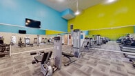 This fitness center is a comprehensively equipped exercise facility designed to support a wide range of workout routines. The space is defined by its striking interior design, featuring bold yellow-green and sky-blue accent walls that create an energizing atmosphere. Full-length mirrors line multiple walls, visually expanding the space and allowing users to monitor their form throughout their workouts.

The cardio section is well-stocked with a row of treadmills, elliptical trainers, stair climbers, and stationary bikes, providing ample options for aerobic exercise. Complementing this is a dedicated strength training area outfitted with a variety of cable and resistance machines suited for full-body conditioning. A free weights station with dumbbells and exercise balls rounds out the strength training offerings.

The facility features high vaulted ceilings with clerestory windows that allow natural light to filter in from above, further brightening the interior alongside pendant and recessed lighting. Ceiling fans support air circulation throughout the space. Multiple wall-mounted flat-screen televisions are positioned throughout the room for entertainment during workouts. The floor is covered in durable, textured carpet tiles that provide both comfort and traction. Overall, this fitness center offers a vibrant, functional, and well-organized environment for a thorough workout experience.