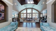 This single-story clubhouse offers an impressive and thoughtfully designed interior centered around a dramatic vaulted ceiling with a distinctive barrel-arch profile and a bold, dark-finished central ceiling detail that draws the eye upward. The grand architectural design is complemented by a large arched window wall that spans the front of the main gathering area, flooding the space with abundant natural light and offering views of the surrounding landscaped greenery and mature trees beyond.

The main lounge area features rich, dark hardwood flooring that anchors the space and contrasts beautifully with the light-toned walls. A generously sized seating arrangement—comprising neutral-toned sofas, patterned accent chairs in soft teal, and a central coffee table atop an area rug—creates an inviting atmosphere ideal for socializing or relaxing. A fireplace anchors one end of the room, adding warmth and a focal point to the space.

Recessed lighting throughout supplements the natural light, ensuring the interior remains bright and welcoming at all hours. The overall ambiance is polished yet comfortable, blending elegant architectural elements with approachable, residential-style furnishings.