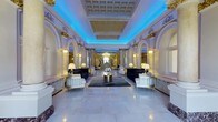 Located at 699 North Broad Street in Philadelphia, PA, this magnificent venue showcases a grand, single-floor hall that exemplifies classical Beaux-Arts architecture at its finest. The expansive main hall commands immediate attention with its soaring coffered ceiling, dramatically illuminated by vibrant blue ambient lighting that casts a striking contrast against the bright white ornamental plasterwork above.

The space is lined with a series of elegant arched alcoves, each framed by intricately carved pilasters and crowned with gilded decorative accents, lending a sense of grandeur and historical richness throughout. Warm wall-mounted sconces are positioned along the arched niches, providing a soft, welcoming glow that complements the bold overhead lighting.

The polished marble flooring reflects the luminous atmosphere, enhancing the sense of openness and refinement. Tastefully arranged seating — including dark velvet sofas and ivory accent chairs — is centered around a gleaming gold circular table, offering an inviting and sophisticated gathering space. A chandelier visible deeper within the hall reinforces the venue's opulent character.

Overall, this space exudes a blend of historic elegance and contemporary ambiance, making it an impressive and versatile setting suited for high-profile events, receptions, or formal gatherings in the heart of Philadelphia.