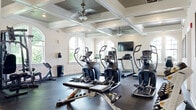 This fitness room offers a thoughtfully designed, single-floor facility that provides a comprehensive workout environment. The main exercise area is defined by its striking coffered ceiling with beadboard insets, ceiling fans, and recessed lighting, lending the space an upscale architectural character uncommon in typical gym settings. Arched windows line the walls, flooding the workout floor with natural light and offering views of the surrounding greenery.

The exercise floor is covered in durable rubber flooring and equipped with a robust selection of cardio machines, including treadmills, ellipticals, and a stationary bike, alongside a cable weight machine, a dumbbell rack with a full range of free weights, and a flat bench. A wall-mounted flat-screen television provides entertainment during workouts. Workout reference charts are displayed on the walls for user guidance.

A dedicated entryway with hardwood flooring and glass-paneled doors connects the facility to the exterior. An adjacent room, finished with the same warm hardwood floors, serves as a children's play area, complete with a wall-mounted TV, activity tables, toys, and a colorful area rug — making the fitness room accommodating for families.