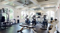 This fitness room offers a thoughtfully designed, single-floor facility that provides a comprehensive workout environment. The main exercise area is defined by its striking coffered ceiling with beadboard insets, ceiling fans, and recessed lighting, lending the space an upscale architectural character uncommon in typical gym settings. Arched windows line the walls, flooding the workout floor with natural light and offering views of the surrounding greenery.

The exercise floor is covered in durable rubber flooring and equipped with a robust selection of cardio machines, including treadmills, ellipticals, and a stationary bike, alongside a cable weight machine, a dumbbell rack with a full range of free weights, and a flat bench. A wall-mounted flat-screen television provides entertainment during workouts. Workout reference charts are displayed on the walls for user guidance.

A dedicated entryway with hardwood flooring and glass-paneled doors connects the facility to the exterior. An adjacent room, finished with the same warm hardwood floors, serves as a children's play area, complete with a wall-mounted TV, activity tables, toys, and a colorful area rug — making the fitness room accommodating for families.