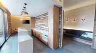 This thoughtfully designed single-floor space combines modern and rustic elements to create a welcoming communal environment. The most prominent design feature is the expansive reclaimed wood accent wall, which runs through the central zone and introduces warm, varied tones of brown and grey throughout the space.

Large-format slate tile flooring in a cool grey hue provides a neutral, durable foundation underfoot. Contrasting the reclaimed wood, white-painted brick walls add texture and brightness, with wall-mounted flat-screen televisions integrated for media display.

A built-in wet bar area anchors one side of the space, featuring warm wood cabinetry, a white countertop, and an undermount sink with a matte black faucet. Full-height cabinetry flanks the bar, maximizing storage while maintaining a clean, cohesive aesthetic.

A cozy recessed lounge nook offers an L-shaped upholstered bench in dark leather, accented with decorative throw pillows. Sculptural wall sconces provide dramatic up-and-down lighting, complemented by subtle LED cove lighting that wraps the perimeter of the nook. Additional seating includes a contemporary lounge chair paired with a bright orange accent table.

A glass door opens to an outdoor patio area, bringing natural light into the space and extending the usable area beyond the interior.