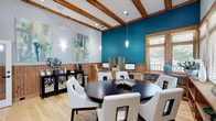 Located at 1140 Trinity Pine Ln in Raleigh, NC, this single-floor community space combines a warm, inviting aesthetic with practical amenities. The interior is immediately distinguished by its bold teal accent walls, which contrast beautifully with crisp white upper walls and rich natural wood wainscoting that lines the lower half throughout. Exposed wood beam ceilings add architectural character and a sense of height to the main area, while pendant lighting and wall sconces provide warm, layered illumination.

The space is thoughtfully arranged into distinct functional zones. A central gathering area features a large, dark wood round table surrounded by white upholstered chairs, making it ideal for meetings or collaborative use. Along the perimeter, built-in wood shelving and countertops house computer workstations, forming a well-equipped business center. A coffee and refreshment station, complete with a single-serve brewer, sits on a stylish black sideboard with decorative glass-panel doors.

A comfortable lounge area offers a tufted white sofa, a glass-top coffee table, and a teal area rug, accented by vibrant artwork and decorative accessories. Multiple windows dressed with wood-framed blinds allow natural light to fill the space while offering views of the surrounding greenery. The light hardwood flooring ties all areas together, creating a cohesive and welcoming environment throughout.