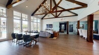 Located at 2325 Nashville Pike in Gallatin, Tennessee, this single-story community clubhouse is a well-appointed gathering space that blends rustic charm with modern design. The main hall commands attention with dramatically vaulted ceilings supported by rich, dark-stained exposed wood beam trusses and an elegant chandelier that anchors the room. Hardwood flooring runs throughout, lending warmth and continuity to the open-plan layout.

Floor-to-ceiling windows and glass walls flood the interior with abundant natural light and offer sweeping views of the outdoor pool and surrounding community grounds. The main lounge area features comfortable seating arranged around a flat-screen TV, creating an inviting space for relaxation or socializing. A dedicated dining area with a solid wood table and upholstered chairs provides additional flexibility for gatherings.

The kitchen is outfitted with white cabinetry, light countertops, and a striking patterned tile backsplash, complemented by pendant lighting and a central island with bar seating. A welcoming reception lounge with wingback chairs and a chandelier pendant light provides a refined waiting area adjacent to the leasing or administrative offices, which are enclosed by frosted glass partition doors. Contemporary artwork throughout adds a curated, polished touch to the overall ambiance.