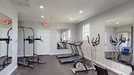 Located at 901 Chalk Level Rd in Durham, NC, this single-floor fitness center is a well-equipped community gym designed to support a comprehensive workout experience. The space is organized into distinct functional zones, accommodating both cardio and strength training activities.

The cardio area features multiple treadmills, elliptical trainers, and stationary bikes arranged in an open layout, allowing for comfortable use of each machine. The strength training section is outfitted with a multi-station cable machine, adjustable weight benches, and a power tower for bodyweight exercises. Large wall-mounted mirrors throughout the space enhance the sense of openness and allow users to monitor their form during workouts.

The gym is finished with durable dark rubber flooring, providing both safety and functionality underfoot. Recessed lighting throughout the ceiling ensures the space is well-lit, while several windows allow natural light to supplement the interior illumination. A dual water fountain station is conveniently positioned near a posted set of gym rules, promoting a clean and orderly environment. A restroom is accessible directly from the main fitness floor. The walls are painted in a clean light gray, contributing to a bright, modern, and welcoming atmosphere suited for daily fitness use.