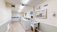 Located at 640 Northern Rd in South Daytona, Florida, this shared laundry facility is a practical, single-floor space designed to serve community residents efficiently. The room features light wood-look tile flooring and neutral two-tone walls with a soft sage wainscoting detail and white trim, creating a clean and welcoming environment. Overhead fluorescent lighting ensures the space is brightly lit throughout.

A row of modern, digital front-load and top-load washers and dryers lines one wall, offering multiple units to accommodate simultaneous use. Stacked dryer units are also present, maximizing the available floor area. A freestanding utility sink provides a convenient handwashing or pre-treatment station, while a built-in fold-down countertop shelf offers a dedicated surface for sorting and folding laundry.

A wooden bench along the opposite wall provides comfortable seating for residents waiting on their laundry. A ceiling fan adds to the comfort of the space. Natural light filters in through plantation-shuttered windows at the far end of the room. A framed bulletin board displays notices and community information, and signage throughout reinforces facility rules. The space is well-maintained, orderly, and thoughtfully arranged to support a smooth and functional laundry experience.