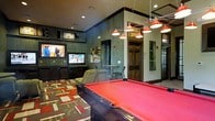 This single-floor recreational space is designed for entertainment and socializing, combining a bold aesthetic with practical amenities. At the heart of the room is a full-size pool table with a vibrant red felt surface, illuminated by striking red pendant lights that hang from a sleek rail-mounted fixture overhead. The flooring features a geometric, multicolored carpet in shades of red, gray, and beige, adding a dynamic visual element to the space.

A built-in media wall along one side of the room houses multiple flat-screen televisions framed by dark cabinetry with ample storage, creating a dedicated lounge area complete with comfortable seating. The walls are dressed in a textured, wood-toned paneling in muted green-gray tones, lending the space a modern yet warm character.

A bistro-style high-top table with bar stools is positioned nearby, offering a casual spot for conversation or gaming. A rustic wooden door with decorative framing adds an architectural accent and connects the space to an adjacent area. Recessed lighting complements the pendant fixtures, ensuring the room is well-lit throughout. Overall, the space strikes a balance between a lively game room atmosphere and a comfortable communal lounge.