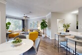 This single-floor residence offers a thoughtfully designed open-concept layout that seamlessly connects the living, dining, and kitchen areas. Rich wood-look flooring runs throughout, complemented by neutral gray walls, crown molding, and textured ceilings that add architectural character. The living room is bathed in natural light from large windows dressed with layered blinds and drapes, and features a ceiling fan, generous wall space for media, and lush greenery that enhances the welcoming ambiance. Adjacent to the living area, the dining space accommodates a round marble-top table and is adorned with a vibrant landscape painting, adding a colorful focal point. The kitchen is equipped with white shaker cabinetry, subway tile backsplash, stainless steel appliances, and a white quartz peninsula with bar seating — ideal for casual dining. A welcoming entryway hallway with decorative wall accents leads guests into the heart of the unit. The bedroom offers a peaceful retreat with natural light filtering through blinds and floor-length curtains, warm wood-toned furnishings, and ample storage. The bathroom presents a clean, modern aesthetic with a white vanity, large mirror, and a shower-tub combination. Overall, the space delivers a bright, contemporary feel with cohesive design details throughout.