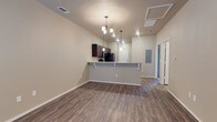 Located at 11206 University in Lubbock, Texas, this single-floor space offers a clean, modern layout designed for comfortable everyday living. The open-concept floor plan connects the living area to the kitchen, creating a seamless and functional flow throughout the main living space. Rich, dark hardwood-style flooring extends across the entirety of the main area, adding warmth and a contemporary aesthetic.

The kitchen is equipped with dark espresso-toned cabinetry and black appliances, and features a raised breakfast bar with built-in corbel supports — ideal for casual dining or additional workspace. A stylish chandelier with multiple light fixtures hangs above, complementing the recessed lighting and providing ample illumination throughout the open space.

The ceilings are notably elevated, contributing to an airy, open feel that enhances the overall sense of spaciousness. Neutral, light-toned walls create a versatile backdrop throughout. A hallway visible beyond the kitchen leads to additional rooms, and an electrical panel is conveniently accessible from the main living area. HVAC vents are integrated into the ceiling, ensuring year-round climate comfort. White trim and door casings provide a crisp, finished look throughout the space.