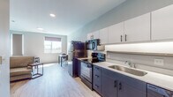 This single-floor unit showcases a clean, contemporary design with a cohesive two-tone color palette of dark navy cabinetry and crisp white surfaces carried throughout the space. The open-concept kitchen is well-appointed with a full suite of black appliances, including a refrigerator, electric range with overhead microwave, and a stainless steel sink. Upper white cabinetry with matte black hardware provides ample storage, while under-cabinet lighting highlights the white countertops below. Light wood-tone luxury vinyl flooring runs seamlessly throughout, enhancing the sense of openness.

The kitchen flows naturally into a dining area and living space, where natural light enters through roller-shade windows. The living area accommodates a sofa, round coffee table, and a low-profile media console, creating a functional yet understated lounge space.

The bedroom features dual windows offering outdoor views and ample natural light, complemented by a practical desk and seating area. The bathroom continues the dark cabinetry and white countertop theme, featuring a single vanity with a large mirror, toilet, and wood-look flooring. A glimpse of an adjacent closet with built-in shelving adds practical storage. Recessed and flush-mount lighting provide consistent illumination throughout.