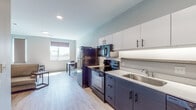 This single-floor unit showcases a clean, contemporary design with a cohesive two-tone color palette of dark navy cabinetry and crisp white surfaces carried throughout the space. The open-concept kitchen is well-appointed with a full suite of black appliances, including a refrigerator, electric range with overhead microwave, and a stainless steel sink. Upper white cabinetry with matte black hardware provides ample storage, while under-cabinet lighting highlights the white countertops below. Light wood-tone luxury vinyl flooring runs seamlessly throughout, enhancing the sense of openness.

The kitchen flows naturally into a dining area and living space, where natural light enters through roller-shade windows. The living area accommodates a sofa, round coffee table, and a low-profile media console, creating a functional yet understated lounge space.

The bedroom features dual windows offering outdoor views and ample natural light, complemented by a practical desk and seating area. The bathroom continues the dark cabinetry and white countertop theme, featuring a single vanity with a large mirror, toilet, and wood-look flooring. A glimpse of an adjacent closet with built-in shelving adds practical storage. Recessed and flush-mount lighting provide consistent illumination throughout.