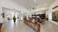 This well-appointed single-floor community amenity center offers a generous footprint thoughtfully divided into distinct functional areas. The main lounge greets visitors with soaring ceilings, light wood-style flooring, and abundant natural light streaming through large windows dressed with clean white blinds. Comfortable seating arrangements, warm decorative accents, and French doors opening to an outdoor pool area create an inviting, resort-like atmosphere. Adjacent to the lounge, a fully equipped communal kitchen features rich dark cabinetry, white countertops, a stainless steel refrigerator, pendant lighting, and an open shelving display wall — ideal for gatherings and social events. A dedicated dining area with farmhouse-style tables and cross-back chairs flows seamlessly from the kitchen, anchored by a charming hand-carved wooden bench as a centerpiece. The fitness center is bright and well-ventilated, furnished with a full cable weight machine, cardio equipment including treadmills and an elliptical, a dumbbell rack, ceiling fans, and mirrored accents. A compact business center rounds out the space, offering multiple computer workstations, a printer, and tasteful décor — providing a quiet, productive environment for residents.