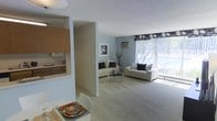 This single-floor space is designed with an open-concept layout that promotes a sense of spaciousness and easy flow between living areas. The living room serves as the centerpiece, featuring plush carpeted flooring, soft blue-grey walls, and large floor-to-ceiling windows dressed with decorative valances that flood the room with natural light. Two cream-toned sofas with accent pillows are arranged around a glass-top coffee table, creating a comfortable and welcoming seating area. A wall-mounted air conditioning unit and a framed art piece add functional and aesthetic touches to the room.

The kitchen is partially open to the main living space via a pass-through window, offering visual connectivity while maintaining a defined cooking zone. Warm wood cabinetry lines the kitchen walls, complementing the neutral color palette throughout. Adjacent to the kitchen, a dining area features a clean white table set with colorful place settings, adding a lively contrast to the otherwise understated décor. The textured ceiling and consistent light carpeting tie the entire space together, giving it a cohesive, calm, and functional ambiance.