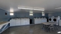 This single-floor laundry facility is designed with practicality and efficiency in mind. The room features two rows of top-load washing machines lined along the back wall, providing ample capacity for multiple users. The machines are uniformly white and neatly arranged, contributing to the clean, organized appearance of the space. Small windows positioned high along the back wall allow for a modest amount of natural light, complementing the bright fluorescent strip lighting mounted overhead that ensures the room is well-illuminated at all times. The walls are finished in a neutral grey tone, and the floor is covered in large-format grey tiles, creating a cohesive, easy-to-maintain aesthetic. A folding utility table is positioned to one side, offering a convenient surface for sorting or folding laundry. A waste bin is centrally placed for easy access. The space is straightforward and functional, with a layout that maximizes usability while maintaining an uncluttered environment suitable for a shared or communal laundry setting.