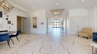 Located at 1085 Goldstream Ave in Victoria, BC, this building lobby presents a polished and welcoming first impression for residents and visitors alike. The space is anchored by expansive marble-effect porcelain tile flooring that runs throughout, lending a refined, upscale aesthetic to the entire area.

The main entrance features floor-to-ceiling glass doors and sidelights framed in sleek black metal, allowing abundant natural light to pour into the corridor and lounge area. The bright, white walls are accentuated by stylish wall sconces and recessed lighting, contributing to a clean and contemporary ambiance.

A comfortable lounge seating area near the entrance includes a plush navy blue sofa, an accent chair, and a marble-topped coffee table, creating an inviting space for residents to gather. Curated wall art adds a decorative touch to this area.

The hallway extends from the entrance toward the elevator bank, which features stainless steel doors, providing convenient vertical access throughout the building. A dedicated mail and parcel room is equipped with a full wall of individual mailboxes, offering secure and organized mail retrieval for all residents.

The space also includes a bathroom for added convenience. A pass-through window provides a view into a kitchenette or utility area, suggesting additional functional support spaces nearby. Overall, this lobby combines practical amenities with elegant design.