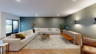 Located at 21 Washington Ave in Carteret, NJ, this generously sized amenity space spans a single floor and seamlessly blends sophisticated interior design with thoughtfully planned outdoor areas. The interior features two distinct gathering zones. The first is a bright, open dining and lounge area finished with herringbone-patterned hardwood flooring, globe pendant lighting, and floor-to-ceiling sliding glass doors that flood the space with natural light and provide direct access to the courtyard. Multiple dining tables with rich green velvet seating chairs are complemented by a well-appointed kitchen with open shelving. Adjacent to the dining space, a relaxed lounge area features deep-toned accent walls, plush neutral sectional sofas, warm ambient lighting, and a striking decorative wall with teal watercolor-patterned wallcovering with banquette seating and modern armchairs. The outdoor courtyard serves as a central gathering hub, featuring a manicured artificial lawn, wide stone-paved walkways, raised planters with ornamental trees and native grasses, and multiple seating vignettes with contemporary outdoor furniture. The surrounding multi-story building facade with balconies frames the courtyard, creating an enclosed, resort-like atmosphere ideal for relaxation and socializing.