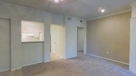 This single-story space showcases an open-concept layout designed for comfortable, functional living. The main living area is finished with wall-to-wall carpet in a neutral tone, complemented by light gray walls and crisp white trim that create a clean, understated aesthetic throughout. Crown molding runs along the ceiling, adding a subtle architectural detail that elevates the overall finish of the space.

A ceiling fan with a light fixture is centrally positioned in the living area, providing both comfort and illumination. Recessed lighting supplements the overhead fixture, ensuring the space is well-lit. HVAC vents are visibly integrated into the ceiling, indicating a central climate control system.

The kitchen is accessible via an open pass-through with a raised breakfast bar counter, offering a convenient connection between the living and kitchen areas. White cabinetry is visible from the living space, contributing to the bright, cohesive color palette. A louvered bi-fold closet door is positioned in the hallway area, likely providing storage access. Additional doorways lead to other areas of the home, suggesting a practical and well-organized floor plan throughout the single level.