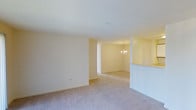 This single-floor space presents a clean, open layout characterized by soft neutral wall tones and plush wall-to-wall carpeting throughout. The main living area is generously proportioned, offering a blank canvas with smooth white ceilings and understated finishes that contribute to a bright, airy atmosphere. The space flows naturally into an adjoining dining area, which is distinguished by an elegant chandelier that serves as a subtle focal point, adding a touch of refinement to the overall design. A partial wall with a pass-through opening separates what appears to be a kitchen area, maintaining visual connectivity between spaces while providing functional division. The consistent use of warm, cream-toned walls throughout creates a cohesive and inviting ambiance. The open sightlines between the living and dining areas enhance the sense of spaciousness, making the layout feel fluid and well-connected. The combination of soft carpeting, neutral palette, and thoughtful lighting fixtures gives the space a comfortable, move-in-ready quality suitable for a variety of furnishing styles and personal preferences.