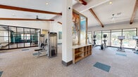 Located at 9270 Eagle Ranch Rd NW in Albuquerque, New Mexico, this fitness center offers a well-appointed, single-floor layout designed for comprehensive exercise and wellness. The space is characterized by soaring vaulted ceilings adorned with rich, exposed wooden beams that lend a warm architectural character to the otherwise modern interior. Large floor-to-ceiling windows and glass-panel walls flood the space with natural light, while ceiling fans provide comfortable air circulation throughout.

The main exercise room is generously sized and equipped with a wide array of fitness machines, including ellipticals, treadmills, stationary bikes, cable and strength machines, and a dedicated free-weight area with a full dumbbell rack. Mirrored walls in the weight section enhance the sense of space. A separate, glass-enclosed studio area with hardwood flooring provides space for group classes or focused training. Patterned, durable flooring with teal accent tiles adds a modern aesthetic touch throughout.

Vibrant, large-scale wall murals and curated artwork contribute an energetic and inspiring ambiance. The entry hallway features double glass doors with direct outdoor views, providing easy access and a welcoming transition into the facility. The overall design balances functionality with a polished, community-oriented aesthetic.