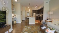 This thoughtfully designed single-floor residence showcases an open-concept floor plan that seamlessly connects the living, dining, and kitchen areas into one cohesive living space. Rich, dark hardwood floors run throughout, grounding the interior with warmth and sophistication.

The kitchen is a standout feature, outfitted with dark espresso cabinetry, stainless steel appliances including a refrigerator, microwave, and range, as well as a striking marble-topped island with seating for multiple bar stools. A decorative pendant light hangs above the island, complementing the track lighting overhead. A classic subway tile backsplash adds texture and visual interest to the cooking area.

The open living area flows naturally from the kitchen and dining space, anchored by a patterned area rug in warm gold and gray tones. Built-in open shelving along one wall provides both functional storage and a place for decorative accents. The high ceilings amplify the sense of space and airiness throughout.

Neutral wall tones in soft white and cream create a clean, versatile backdrop that enhances the natural light filtering into the space. The overall ambiance is modern yet inviting, with a careful balance of dark and light elements that give the home a refined, well-appointed character.
