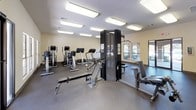 This fitness center offers a comprehensive workout environment designed to support a variety of exercise routines. The single-level space is laid out to maximize usability, with durable rubber flooring throughout that provides cushioning and traction for safe movement during exercise.

The cardio area is well-stocked with multiple treadmills, elliptical trainers, and a stationary bike, providing ample options for aerobic training. Strength training needs are addressed by a multi-station cable machine, along with several weight benches positioned throughout the space to accommodate free weight and resistance exercises.

Large wall-mounted mirrors on one side of the room visually expand the space while allowing users to monitor their form. Recessed fluorescent panel lighting illuminates the entire room evenly from above, ensuring excellent visibility at any time of day. Multiple windows along the exterior walls invite natural light and offer views of the surrounding outdoor environment, contributing to an open and energizing atmosphere.

The space is accessed through glass-panel doors and features clearly marked emergency exit signage. The neutral, light-toned walls and clean layout create a functional and welcoming environment suited for daily fitness activity.