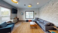 Located at 1001 Toms Creek Rd in Blacksburg, Virginia, this single-floor space is a well-appointed community lounge designed with both comfort and functionality in mind. The main living area is anchored by a generous sectional sofa in cool gray, set atop a woven jute area rug and paired with a solid wood coffee table. Playful touches — including colorful pouf ottomans in orange and charcoal and a bold red metal bar stool — add personality to the space. Two distinct accent walls define the room's character: one features a textured cloud-motif wallcovering, while another showcases a dark, linear patterned wallpaper, creating visual depth and contrast against the crisp white walls. Light wood-look flooring runs throughout, complementing the airy atmosphere. Natural light flows in through multiple windows fitted with clean white blinds, offering views of the surrounding neighborhood. Wall-mounted flat-screen TVs serve both seating areas, and a media console provides additional storage. Adjacent to the lounge, a fully equipped kitchenette features rich dark wood cabinetry, granite countertops, a stainless steel refrigerator, microwave, and a double sink — all finished with light wood flooring that ties seamlessly into the main space. Stylish ceiling fixtures with gold interior accents add a warm, contemporary glow throughout.