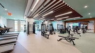 Located at 12 Church Street in New Rochelle, NY, this professionally designed fitness center offers an expansive, single-floor layout that accommodates a full spectrum of workout equipment in a thoughtfully organized space. The interior is defined by a striking ceiling treatment featuring parallel wood slat baffles interspersed with linear LED lighting, lending a warm yet contemporary aesthetic throughout the main exercise area. Rubber flooring in neutral and green tones provides durability and comfort underfoot across the various workout zones. Large floor-to-ceiling windows along the perimeter invite abundant natural light and offer views of the surrounding streetscape, creating an open and energizing atmosphere. The strength training area is well-equipped with cable machines, weight benches, a power rack with Olympic plates, and a comprehensive dumbbell rack. A dedicated cardio zone houses treadmills, ellipticals, stationary bikes, and rowing machines. A separate, glass-enclosed studio area with wood flooring provides an intimate setting for group classes or focused training sessions. Recessed and accent lighting, green-painted ceiling accents, and large mirrors enhance the sense of space throughout. The facility presents a polished, high-end environment suited for a wide range of fitness activities.