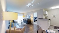 This single-floor residence offers a seamless open-concept layout that effortlessly connects the living, dining, and kitchen areas, creating an inviting and functional living environment. The living room is generously proportioned, featuring rich hardwood-style flooring, neutral gray walls, and large sliding glass doors that bathe the space in abundant natural light. Thoughtfully arranged seating, including a plush sofa and accent chairs, is anchored by a glass coffee table atop a layered area rug, lending the room a polished yet comfortable feel. Decorative framed botanical artwork adds a refined, curated touch to the walls. Adjacent to the living area, the kitchen is visible over a raised countertop bar, offering a practical and social pass-through design with clean white cabinetry and modern finishes. The bedroom, accessed through an open doorway, is a calm and restful retreat, highlighted by a well-dressed bed with coordinated blue bedding and ample space to move comfortably. Throughout the residence, the consistent use of warm wood-toned flooring, soft neutral tones, and contemporary furnishings creates a cohesive and welcoming ambiance that is both modern and livable.