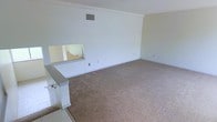 This single-floor space presents a straightforward and functional layout centered around a generously sized main living area. The room is finished with wall-to-wall beige carpeting that extends throughout, providing a neutral and practical foundation for a variety of furnishing arrangements. The walls are painted in a clean, crisp white, contributing to a bright and open atmosphere.

A notable architectural detail is the pass-through window integrated into the partial wall along one side of the room. This opening connects the living area to an adjacent space—likely a kitchen or utility area—enhancing the sense of connectivity between rooms while maintaining a degree of separation. The partial wall itself features a smooth white finish and appears to incorporate a small countertop ledge at the pass-through.

Overhead, a ceiling-mounted HVAC vent is visible, indicating a central air and heating system. The room includes multiple electrical outlets positioned at standard intervals along the baseboards, offering practical placement for furniture and electronics. The overall ambiance is clean, minimal, and move-in ready, making it a versatile space suitable for various uses.