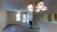 This single-story space centers around a comfortable living area distinguished by a white-mantled fireplace with decorative diagonal paneling above, serving as a natural focal point. The room is finished with wall-to-wall carpet and neutral-toned walls, creating a warm, understated atmosphere. Overhead, a multi-bulb chandelier with bronze detailing provides ambient lighting, complemented by a ceiling fan that enhances air circulation and adds a casual, practical touch. Two windows along the exterior wall allow natural light to filter into the space, offering views of surrounding greenery. A white-paneled door provides direct exterior access, adding convenience. The textured popcorn ceiling is consistent throughout, lending a classic residential character. An adjacent area, visible through an open passageway, suggests additional functional space such as a kitchen or utility area. The overall layout feels open and connected, with the living area flowing naturally toward surrounding rooms. The combination of the fireplace, carpeted floors, and layered lighting gives the space a relaxed, livable quality suited for everyday comfort.