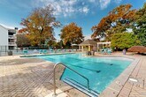 Located in the Brookhaven neighborhood of Atlanta, this expansive outdoor amenity area at 100 Lenox Park Circle NE offers a resort-style retreat for residents. The centerpiece of the space is a pair of generously sized swimming pools with crystal-clear blue water, lap lanes, and wide pool decks finished in a combination of stamped brick pavers and concrete surrounds. Multiple entry points with stainless steel handrails provide easy access to both pools.

The pool deck is well-appointed with teal lounge chairs and umbrella-shaded seating areas, offering ample space for sunbathing and relaxation. A charming covered gazebo with a hipped roof serves as a central gathering point, complemented by a white pergola structure that houses an outdoor grilling station and additional dining furniture — ideal for outdoor entertaining.

A fire pit area adds a warm, social focal point within the landscaped grounds. The entire area is framed by mature trees displaying vibrant autumn foliage in rich shades of gold, orange, and red, creating a picturesque and tranquil atmosphere. Lush ornamental plantings and large decorative planters enhance the natural beauty of the surroundings. A secure perimeter fence encloses the space, and the adjacent clubhouse building provides convenient access to additional amenities.