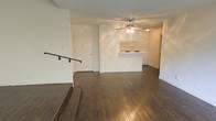 This single-floor residence offers a welcoming and functional layout centered around an open-concept living space. Rich, dark hardwood flooring extends throughout, adding warmth and contrast to the bright, white-painted walls and textured ceiling. A ceiling fan with an integrated light fixture anchors the main living area, providing both comfort and ambient illumination.

The kitchen is positioned at the far end of the open living space and features crisp white cabinetry and a peninsula counter that serves as a natural divider between the kitchen and living areas. The clean, minimalist design of the kitchen complements the overall neutral palette of the home.

A staircase with a classic black metal railing is visible at the front of the space, adding a subtle architectural detail and suggesting a split-level or raised entry element. The entry door, centrally located at the rear of the living area, is framed by white trim and contributes to the bright, airy feel of the space.

Overall, the residence presents a clean, open, and practical interior with consistent finishes throughout. The combination of dark hardwood flooring, white walls, and an open floor plan creates a balanced and versatile living environment suitable for a variety of furnishing styles.
