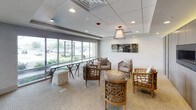 This thoughtfully designed single-floor space functions as an elegant lounge or common area, blending contemporary aesthetics with comfortable, inviting furnishings. The layout is divided into two distinct zones that flow seamlessly into one another. The interior lounge area features a full-wall panel of weathered grey wood cladding housing a sleek, recessed fireplace, providing a warm and refined focal point. A large decorative mirror with a dark, antiqued finish adorns the adjacent white wall, adding depth and artistic character. Woven rattan chairs with white cushions and organic-form wooden coffee tables contribute to a relaxed yet upscale atmosphere. Recessed lighting and subtle cove lighting along the wood wall panels create a layered, ambient glow. The second zone opens toward an expansive wall of floor-to-ceiling windows, flooding the space with abundant natural light and offering views of the surrounding outdoor landscape. A narrow console table with angular legs runs along the window line, complemented by additional seating arrangements. A statement pendant lantern fixture anchors both zones, tying together the overall design. Neutral carpet throughout and a clean drop ceiling with recessed lighting complete the polished, professional ambiance of this versatile space.