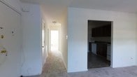 This single-floor residence offers a clean, straightforward layout designed for comfortable everyday living. A central hallway serves as the spine of the home, finished with light carpet underfoot and crisp white walls that reflect natural light filtering in from the far end, creating an open and airy feel throughout. Flush-mount ceiling lighting provides consistent illumination along the corridor, which connects the various areas of the home with ease. To one side of the hallway, a doorway opens into a dedicated kitchen space, where dark-toned cabinetry provides a rich contrast against the lighter surroundings. The kitchen features what appears to be a stainless steel dishwasher and a dark countertop surface, offering a functional workspace with a cohesive, contemporary aesthetic. The flooring in the kitchen transitions to a hard surface, clearly delineating it from the carpeted living spaces. Doors along the hallway suggest additional rooms branching off the main corridor, contributing to a well-organized floor plan. The overall ambiance of the space is neutral and move-in ready, with a clean palette of whites and soft tones that provide a versatile backdrop suitable for a variety of interior styles.