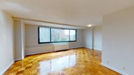 This single-floor residence offers a well-organized layout with distinct living, kitchen, and bathroom areas. The living space is the focal point of the unit, showcasing polished parquet flooring arranged in a classic checkerboard pattern that adds warmth and character throughout. A large picture window spans much of the exterior wall, flooding the room with natural light and framing views of the surrounding urban skyline and greenery below. A built-in radiator beneath the window provides in-unit heating. The clean white walls and smooth ceilings contribute to an open, airy atmosphere.

The kitchen is a compact, efficiently arranged galley-style space featuring white cabinetry, dark granite-style countertops, a stainless steel sink, and a full-size white refrigerator. The floor transitions to durable tile, practical for a food preparation area.

The bathroom is finished with classic white square tiles accented by a green tile border near the ceiling, lending a retro aesthetic. It includes a full-sized soaking tub with an overhead shower, built-in wall niches, and chrome fixtures. Additional rooms visible in the floor plan include a closet and utility areas, providing practical storage solutions. Overall, the space presents a clean, functional, and well-lit urban living environment.
