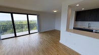 This single-floor space is designed with an open-concept layout that maximizes both functionality and natural light. The living area is anchored by warm-toned hardwood flooring that extends throughout, lending a clean and cohesive aesthetic to the interior. Neutral white walls provide a versatile backdrop, complementing the contemporary finishes found across the space.

A large sliding glass door serves as a prominent architectural feature, opening directly onto a private balcony enclosed by a sleek black metal railing. The balcony offers expansive views of a lush, tree-lined landscape, bringing a strong connection to the outdoors into the living area. The floor-to-ceiling glass allows abundant natural light to fill the room throughout the day.

Adjacent to the living area, the kitchen is partially visible through an open pass-through, featuring dark cabinetry and a light-toned countertop. The layout creates a natural flow between cooking and living spaces while maintaining a sense of openness. Recessed and flush-mounted ceiling lighting supplements natural light, ensuring the interior remains well-lit at all hours. Overall, the space presents a modern, airy, and well-appointed environment.