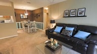 This single-floor space showcases an open-concept layout that seamlessly connects the living and dining areas, creating a cohesive and functional flow throughout. The living room is anchored by a dark leather sofa adorned with blue and patterned accent pillows, complemented by a glass-top coffee table styled with decorative candles and stones. A tall floor lamp provides ambient lighting, while a wall-mounted shelf displays framed artwork, adding a curated touch to the space. The neutral carpeted flooring runs throughout, contributing to a warm and inviting atmosphere. A half-wall with teal-striped detailing serves as an architectural divider between the living and dining areas, adding a distinctive design element. The dining area features a glass-top table surrounded by white ornate chairs beneath a modern pendant light, set against a rich brown accent wall with decorative wall pieces. The overall ambiance is warm and transitional, blending dark and light tones with carefully chosen accessories to create a comfortable, well-styled interior.