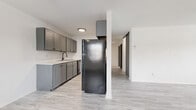 This single-floor unit presents a thoughtfully updated interior with a consistent neutral palette and modern finishes throughout. The open living area is spacious and bright, featuring light wood-style luxury vinyl flooring, white walls, a ceiling fan, and a wall-mounted air conditioning unit. A baseboard heating system runs along the perimeter, and multiple windows dressed with vertical blinds allow natural light to filter in.

The kitchen is positioned adjacent to the living area and is outfitted with gray shaker-style cabinetry on both upper and lower sections, a marble-look countertop, a single-basin sink with a modern faucet, and black appliances including a full-size refrigerator. A structural column separates the kitchen from the living space, adding architectural definition.

The bedrooms are carpeted in a neutral tone and each feature windows with vertical blinds and baseboard heaters, providing a quiet, comfortable atmosphere. At least one bedroom includes a walk-in closet with wire shelving for organized storage.

The bathroom is well-appointed with a gray vanity, marble-look countertop, a wall-mounted mirror, and a tub-shower combination, maintaining the cohesive modern aesthetic found throughout the unit.
