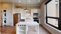 Located at 207 14th Ave SW in Rochester, Minnesota, this single-floor unit offers a clean, modern aesthetic throughout. The kitchen serves as a standout feature, outfitted with white shaker-style cabinetry, quartz countertops, a white subway tile backsplash, and a full suite of stainless steel appliances including a refrigerator, electric range, over-the-range microwave, and dishwasher. A white center island with built-in shelving and a pendant light above provides additional prep space and storage. Large black-framed windows admit generous natural light and offer views of the surrounding Rochester cityscape. Light wood-tone luxury vinyl plank flooring runs throughout the unit, adding warmth and cohesion to the space. The living area is open and versatile, featuring neutral warm-toned walls and recessed lighting. The bathroom is thoughtfully designed with warm wood-tone flooring, white cabinetry with matte black hardware, a full tub-shower combo, and clean white wall finishes. Recessed lighting and matte black door hardware are consistent design details found throughout, lending the space a polished, contemporary feel. The overall layout is efficient and well-lit, making it a functional and attractive urban living space.
