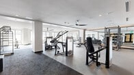 This expansive fitness center, located in Oakland, CA (94608), is a thoughtfully designed, single-floor facility offering a comprehensive workout environment. The space is organized into distinct zones, each tailored to a specific type of training, creating a functional and efficient layout for users of all fitness levels.

The strength training area features commercial-grade Cybex resistance machines, a fully loaded free weights section with a tiered dumbbell rack, and power racks with bench press stations, all set against floor-to-ceiling mirrors that enhance the sense of space. The flooring transitions between durable rubber and dark carpet, clearly delineating training zones.

The cardio area is positioned along a wall of large windows, allowing generous natural light to fill the space and providing outdoor views during workouts. Equipment here includes treadmills, ellipticals, and air-resistance rowing machines. A dedicated functional training zone accommodates a cable climbing machine and open floor space for stretching or bodyweight exercises.

Throughout the facility, ceiling fans maintain air circulation, and recessed lighting complements the abundant daylight. Black-framed glass doors provide a sleek, modern entryway, while framed athletic photography adorns the walls. Warm wood cabinetry and built-in storage along one wall add a refined, upscale touch to the overall aesthetic.