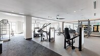 This expansive fitness center, located in Oakland, CA (94608), is a thoughtfully designed, single-floor facility offering a comprehensive workout environment. The space is organized into distinct zones, each tailored to a specific type of training, creating a functional and efficient layout for users of all fitness levels.

The strength training area features commercial-grade Cybex resistance machines, a fully loaded free weights section with a tiered dumbbell rack, and power racks with bench press stations, all set against floor-to-ceiling mirrors that enhance the sense of space. The flooring transitions between durable rubber and dark carpet, clearly delineating training zones.

The cardio area is positioned along a wall of large windows, allowing generous natural light to fill the space and providing outdoor views during workouts. Equipment here includes treadmills, ellipticals, and air-resistance rowing machines. A dedicated functional training zone accommodates a cable climbing machine and open floor space for stretching or bodyweight exercises.

Throughout the facility, ceiling fans maintain air circulation, and recessed lighting complements the abundant daylight. Black-framed glass doors provide a sleek, modern entryway, while framed athletic photography adorns the walls. Warm wood cabinetry and built-in storage along one wall add a refined, upscale touch to the overall aesthetic.